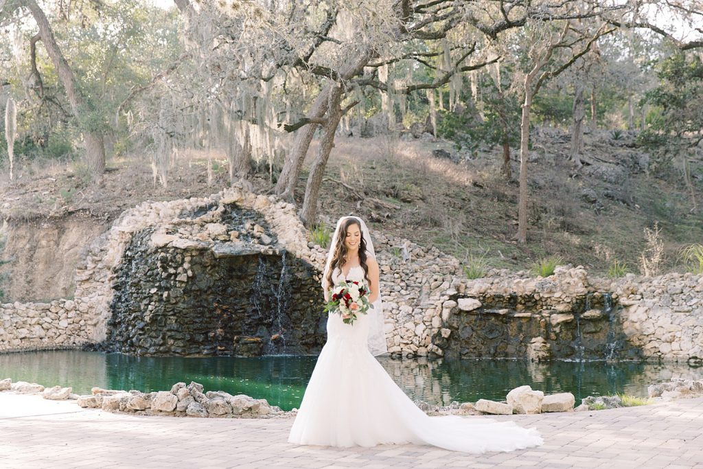 bride looking at her bouquet while standing in front of a waterfall for her Bridal Portraits at Hayes Hollow in Spring Branch, TX with Monica Roberts Photography www.monicaroberts.com